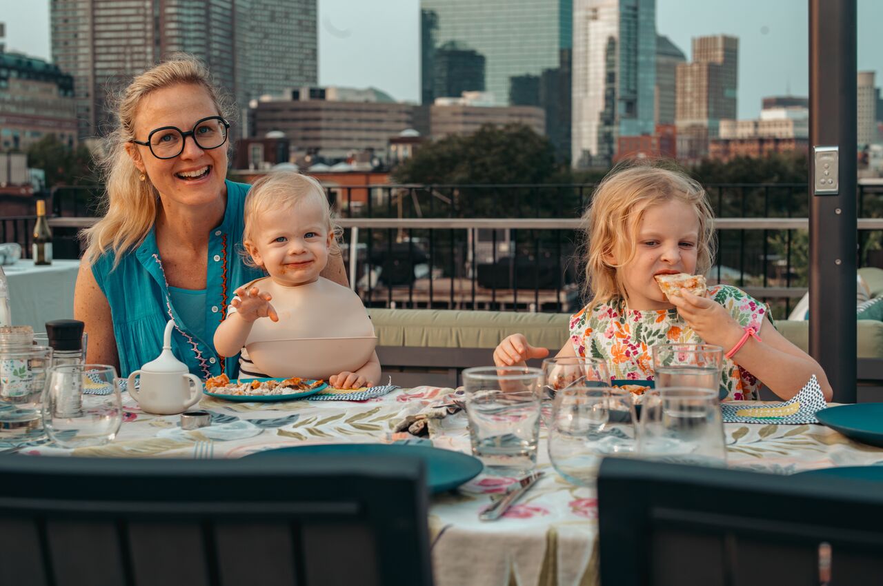 A woman sits at a table with two young children, one smiling with food and the other eating a slice of pizza.