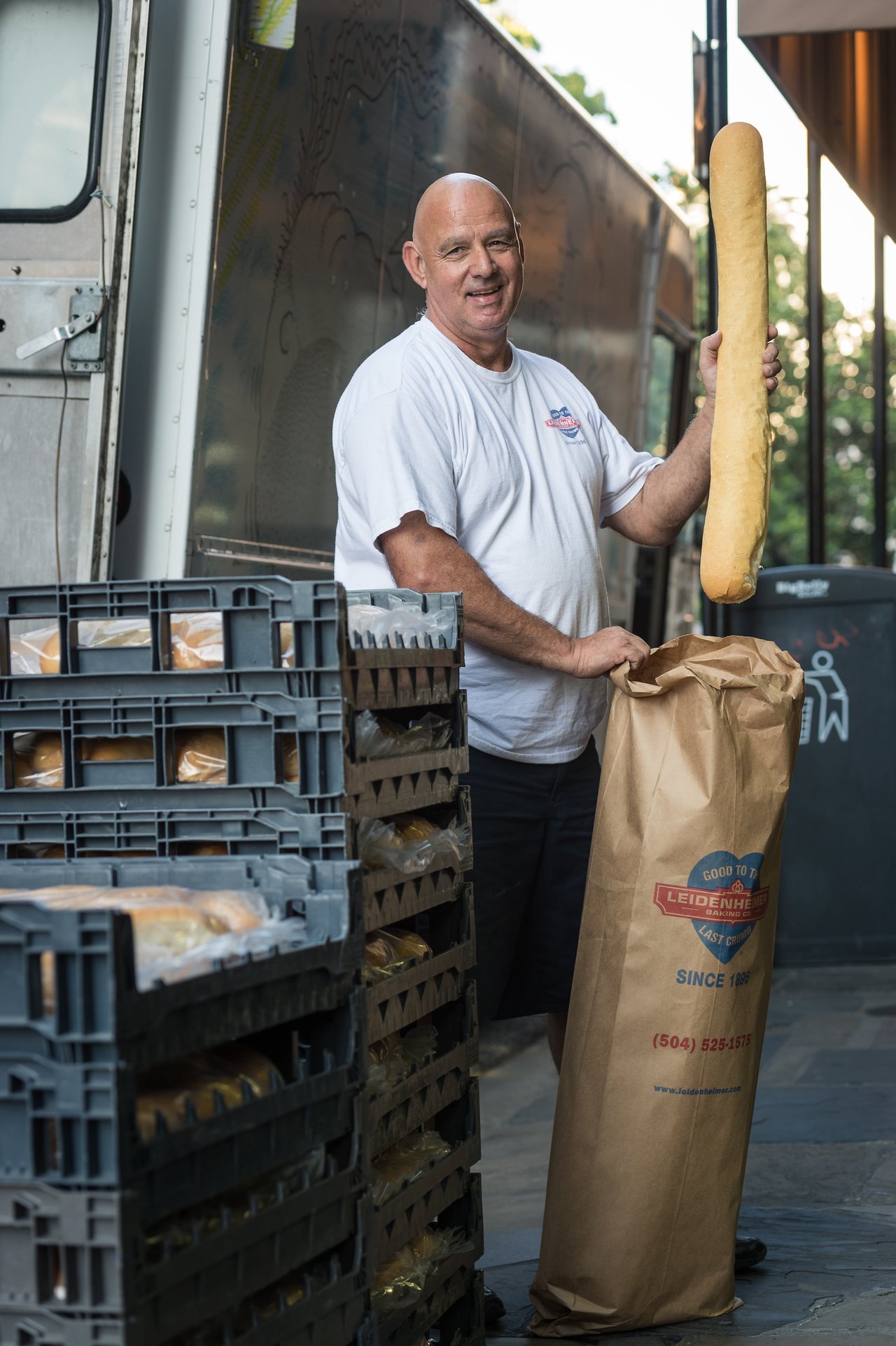 A man in a white shirt holds a large baguette while standing next to stacked bread crates and a truck.
