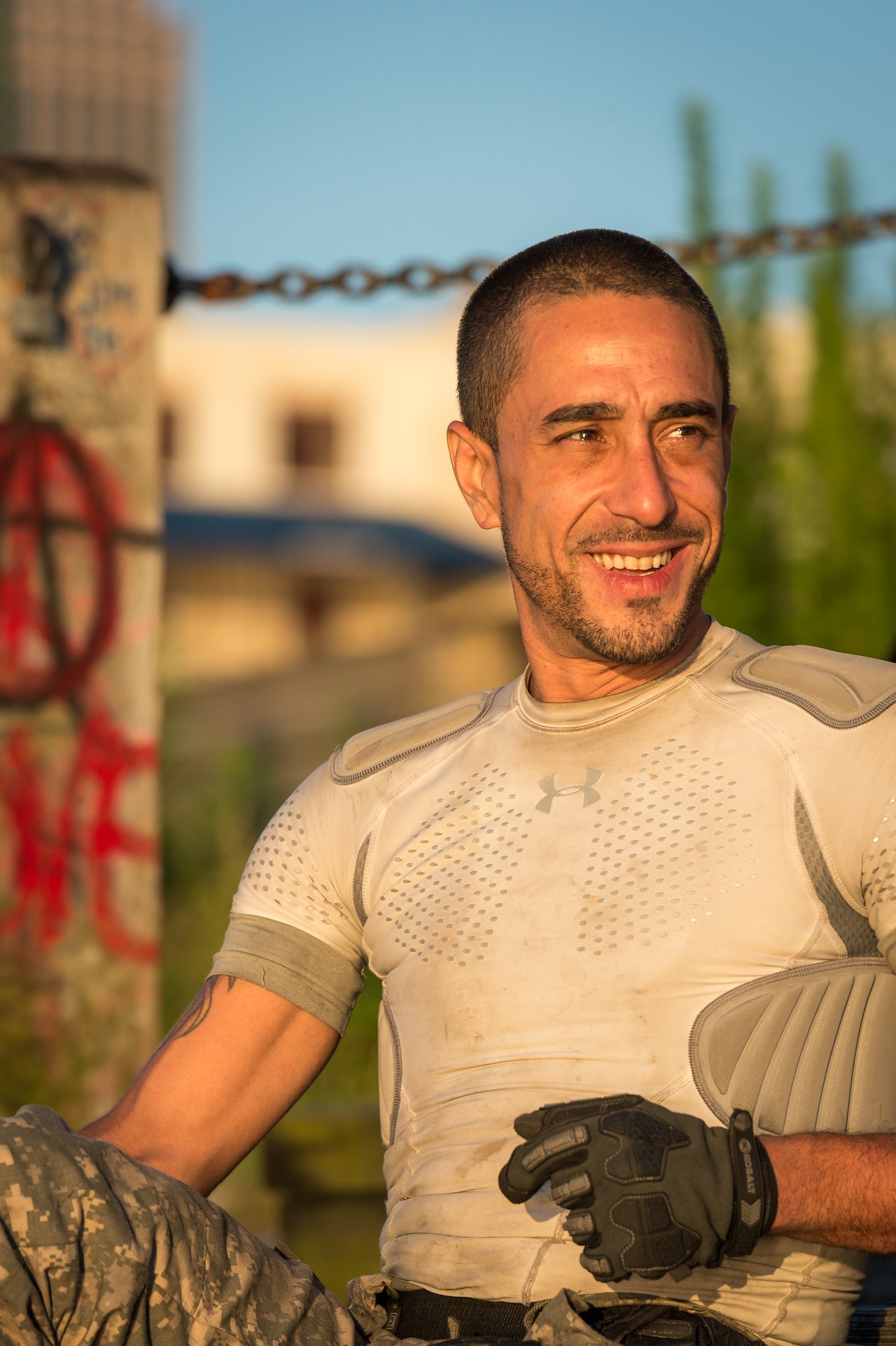 A man in protective gear and gloves sits outdoors, smiling in warm sunlight.