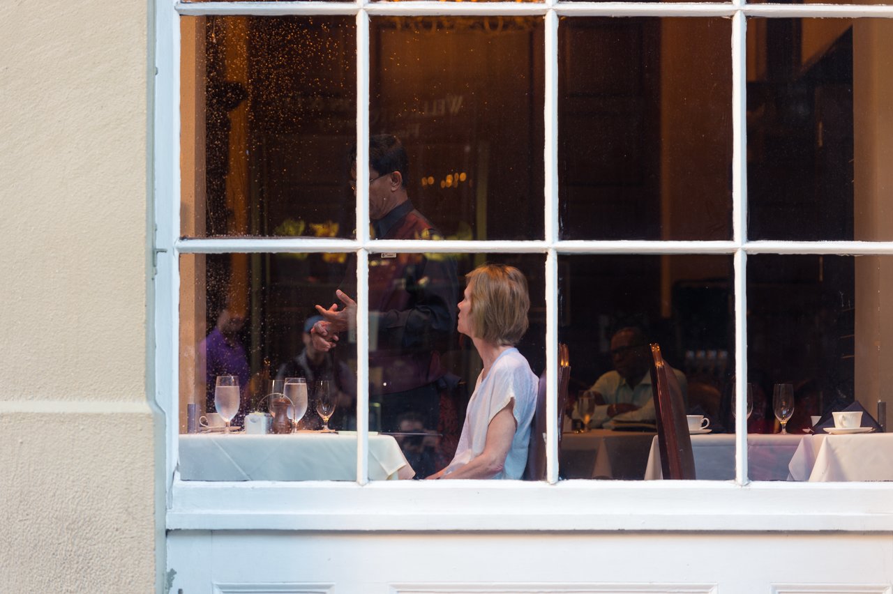 A woman sits at a table inside a hotel restaurant, looking away, while a waiter interacts with another guest.