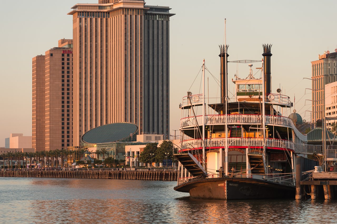 The Steamboat Natchez is docked along the waterfront, with tall buildings in the background during sunset.