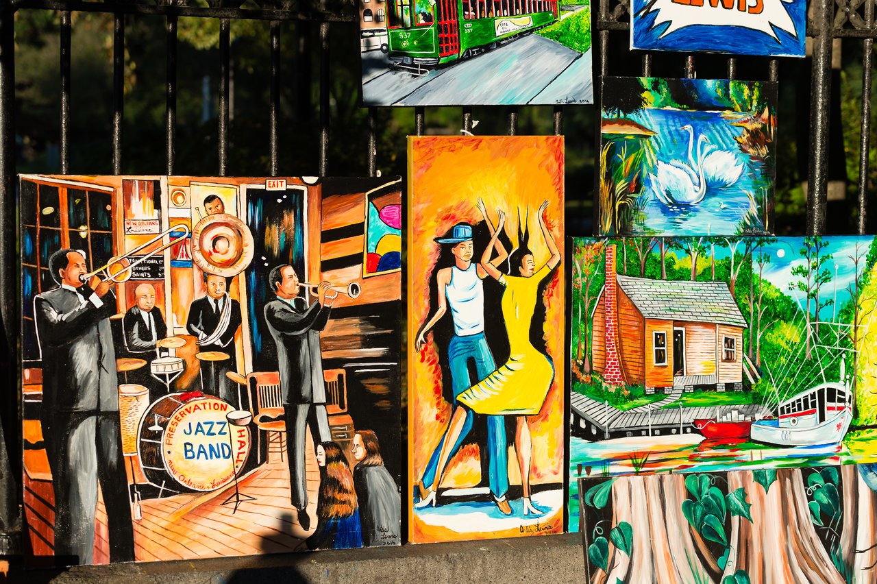 Colorful paintings displayed on a fence, featuring jazz musicians, dancing couples, swans, a streetcar, and a riverside cabin.