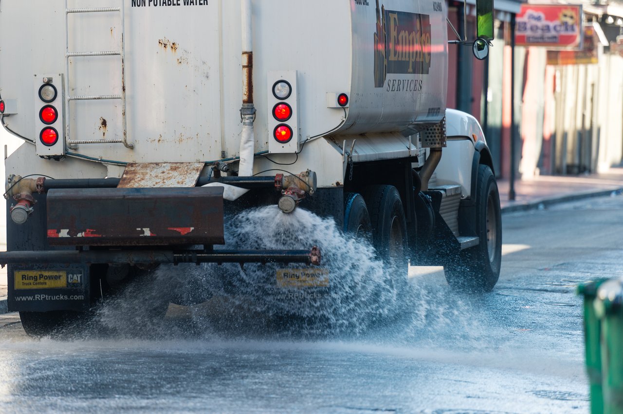 A street cleaning truck sprays water onto the road, washing the pavement in an urban area.
