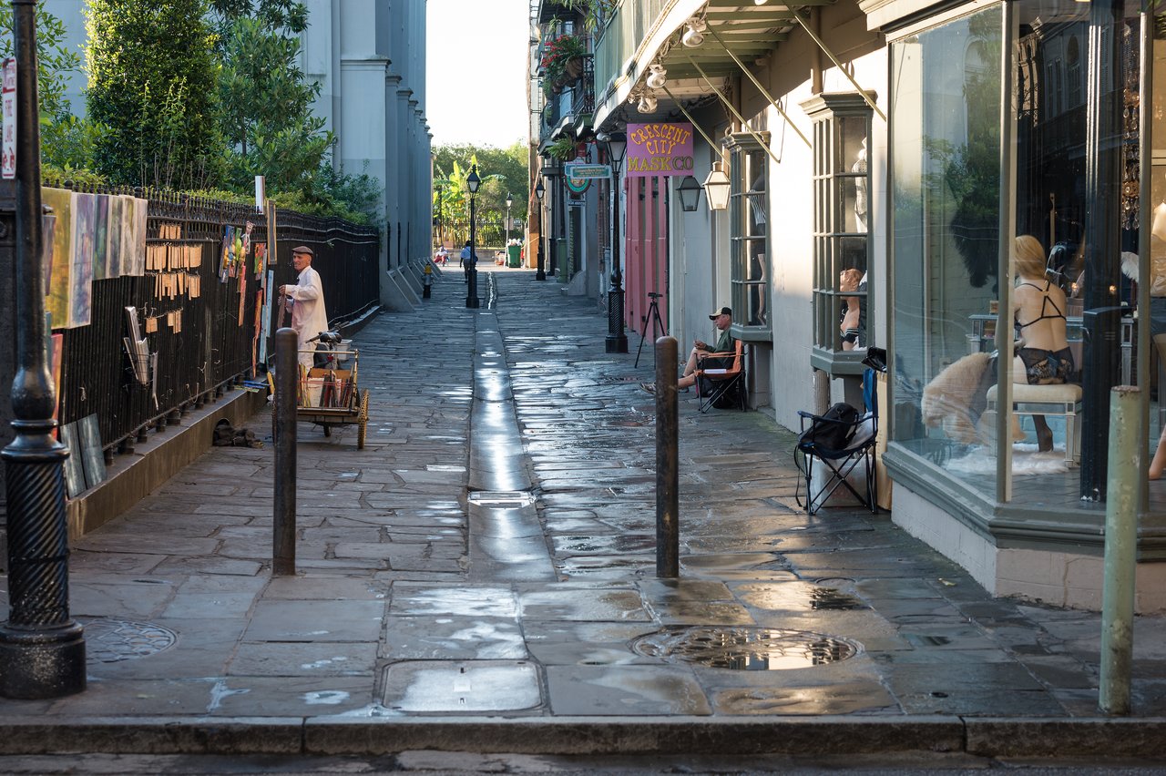 An artist arranges paintings on a fence while people sit outside a shop on a narrow, wet street.