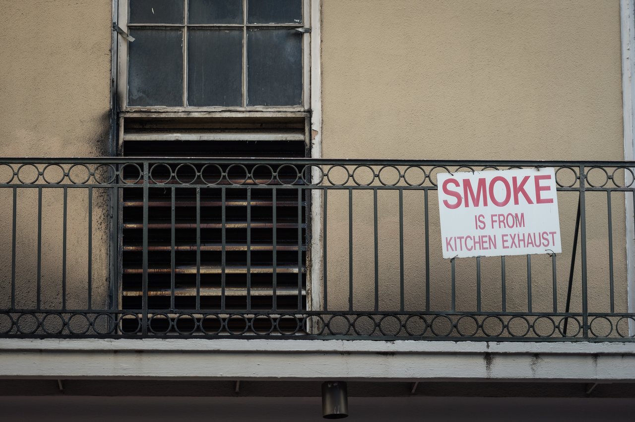 A metal balcony railing with a sign that reads, "SMOKE IS FROM KITCHEN EXHAUST", near a vented window.