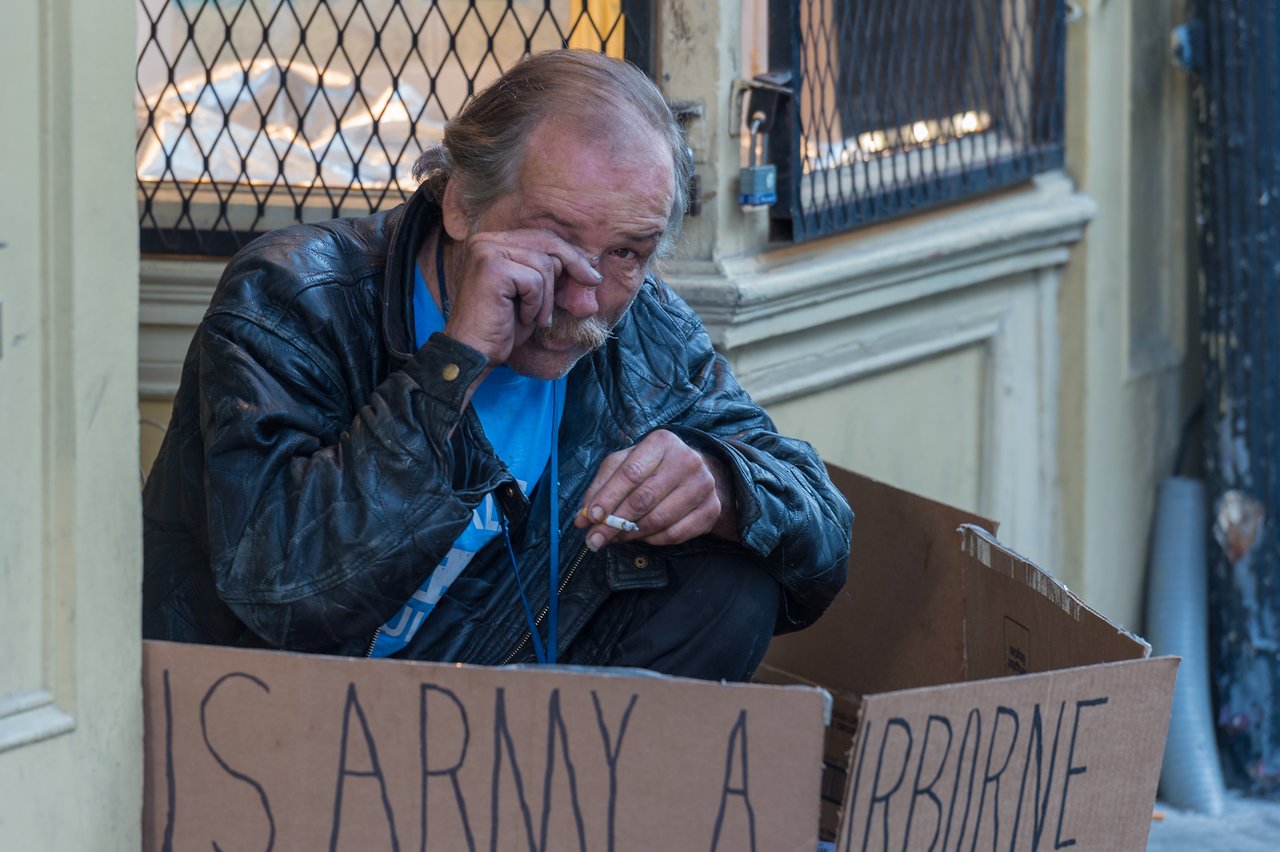 A man in a leather jacket sits on the sidewalk with cardboard signs, wiping his eye and holding a cigarette.