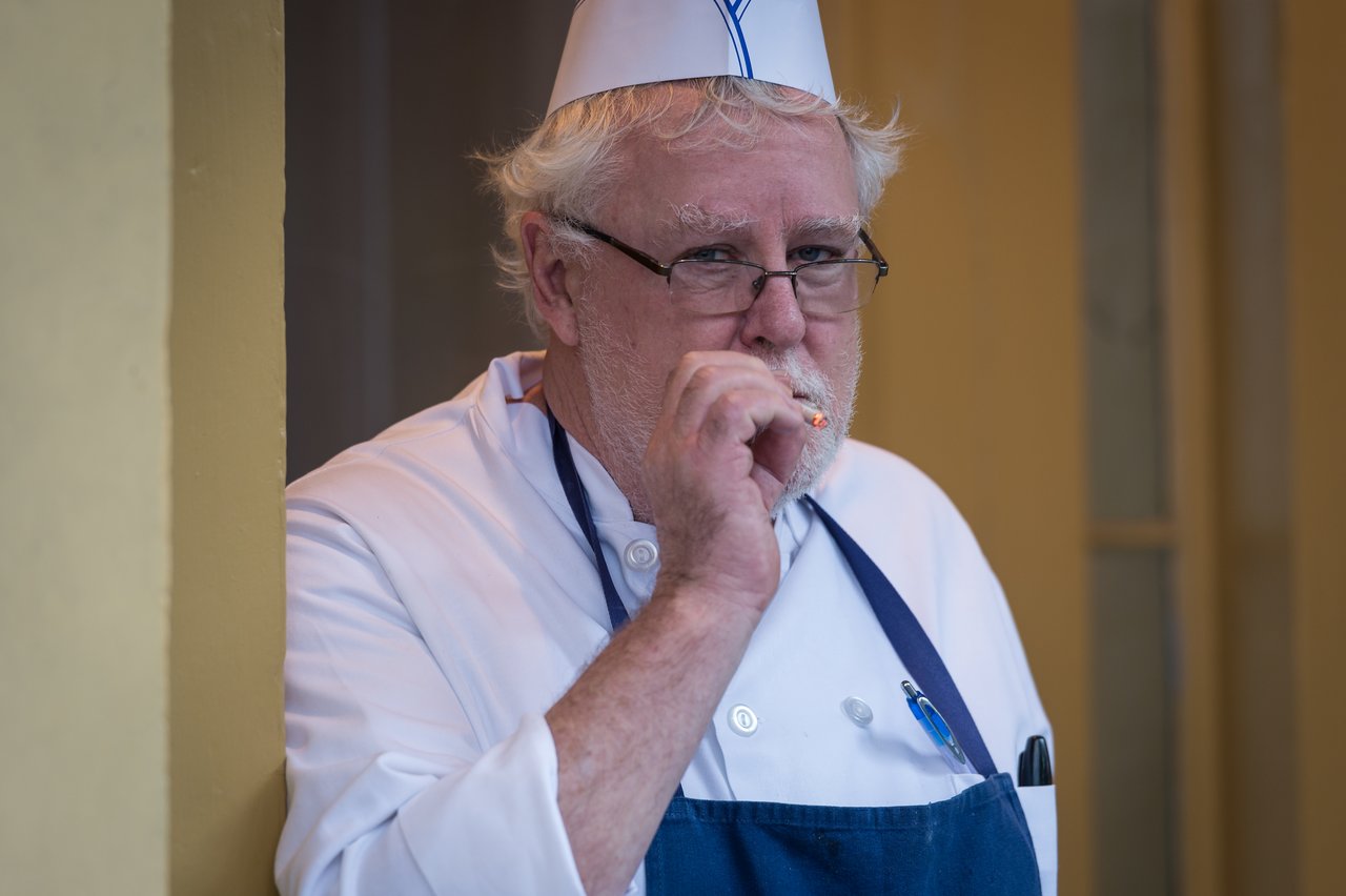 A chef in a white uniform and blue apron smokes a cigarette while standing outside.