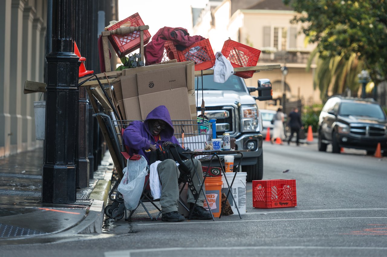 A person in a purple hoodie sits on a chair beside a cart filled with belongings on a city sidewalk.