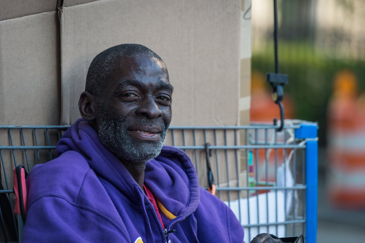 A man in a purple hoodie sits next to a shopping cart filled with belongings, smiling at the camera.