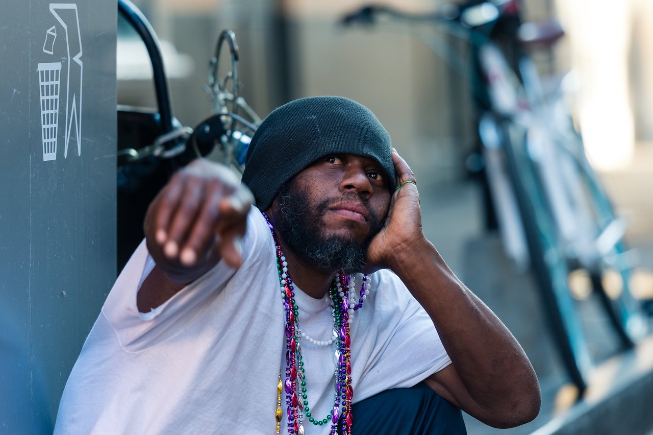 A man wearing a beanie and colorful beads sits on the street, resting his head and pointing forward.