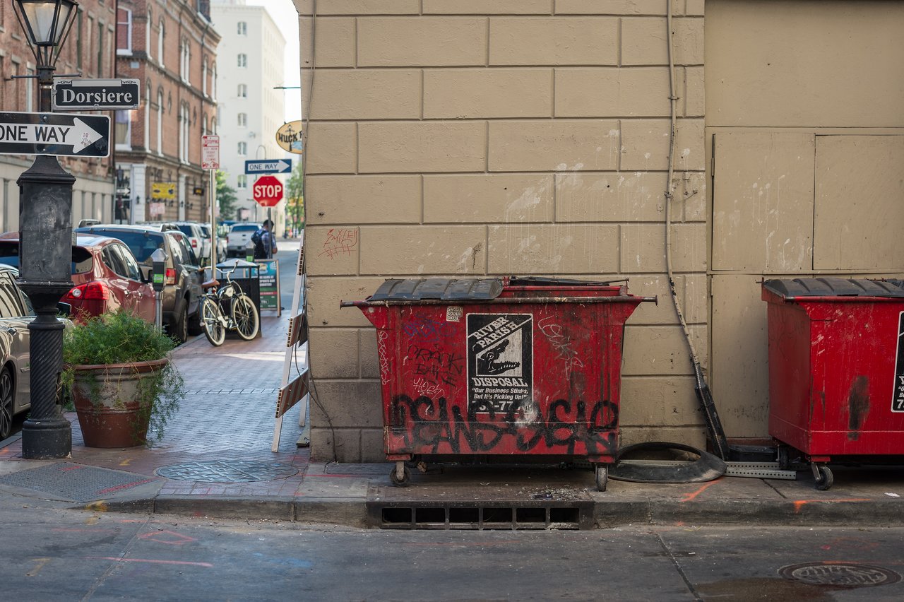 Two red dumpsters with graffiti sit against a beige wall, while parked cars and a bicycle line the street.