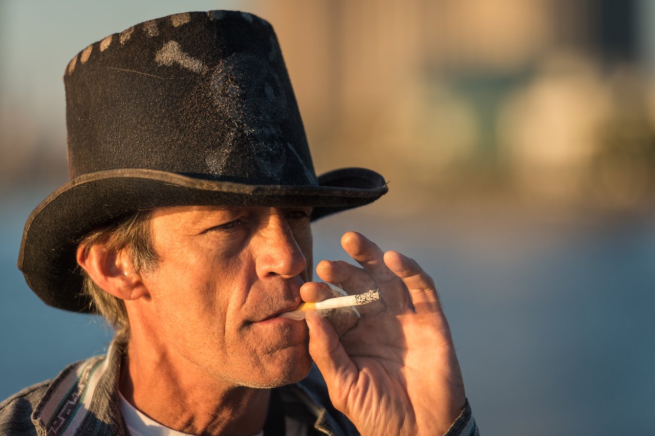 A man wearing a black hat smokes a cigarette outdoors with blurred buildings in the background.