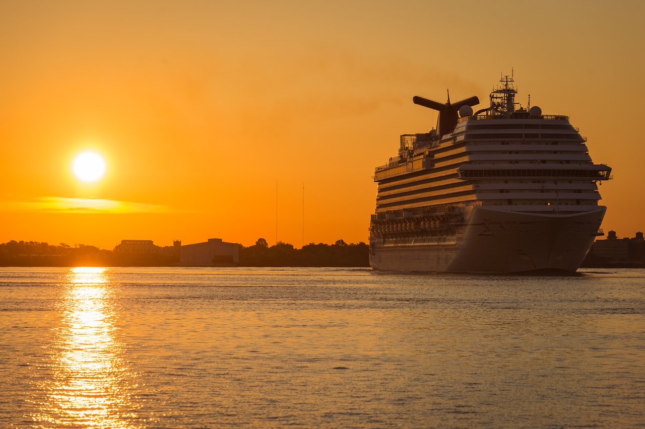 A large cruise ship moves through the water at sunrise, with the sun reflecting on the surface.