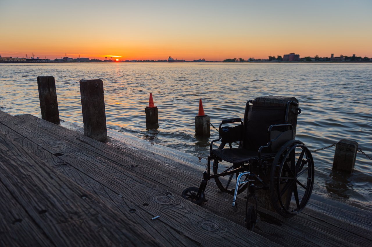 A wheelchair sits on a wooden dock overlooking the water as the sun rises in the distance.