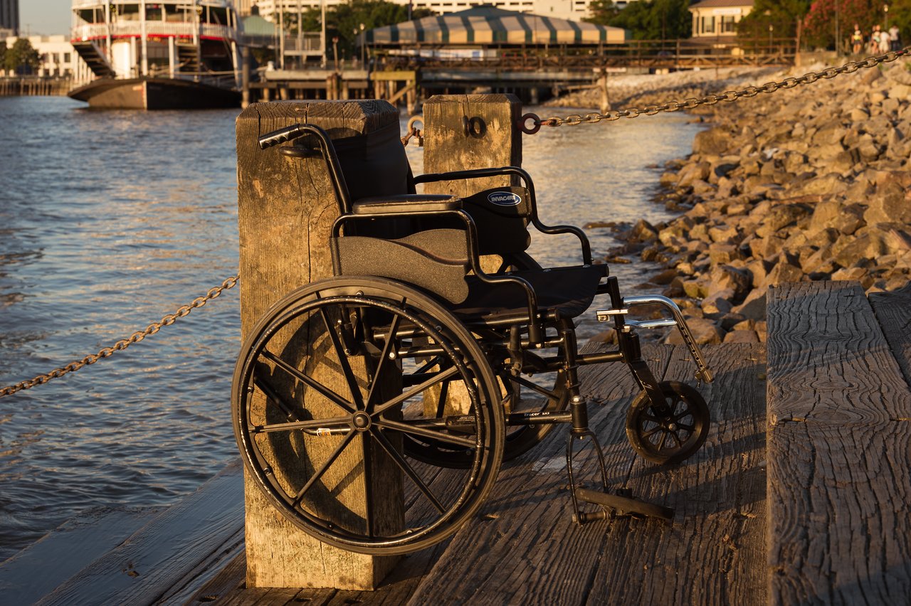 An empty wheelchair is positioned near a wooden dock by the water, illuminated by the warm morning sunlight.