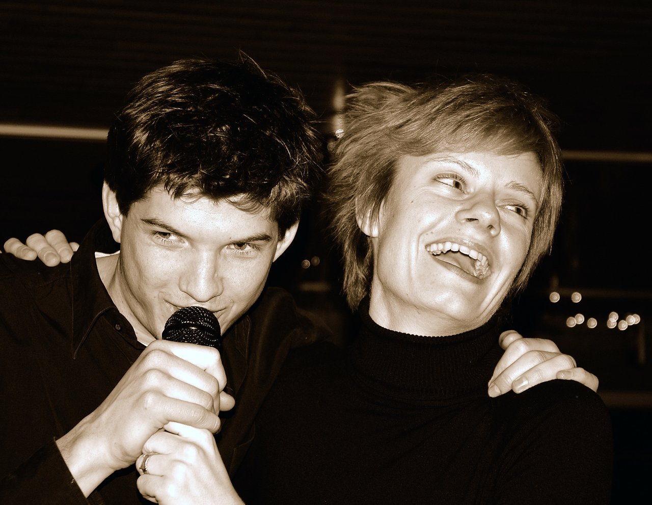 A man sings into a microphone while a woman laughs beside him, both enjoying a karaoke moment.