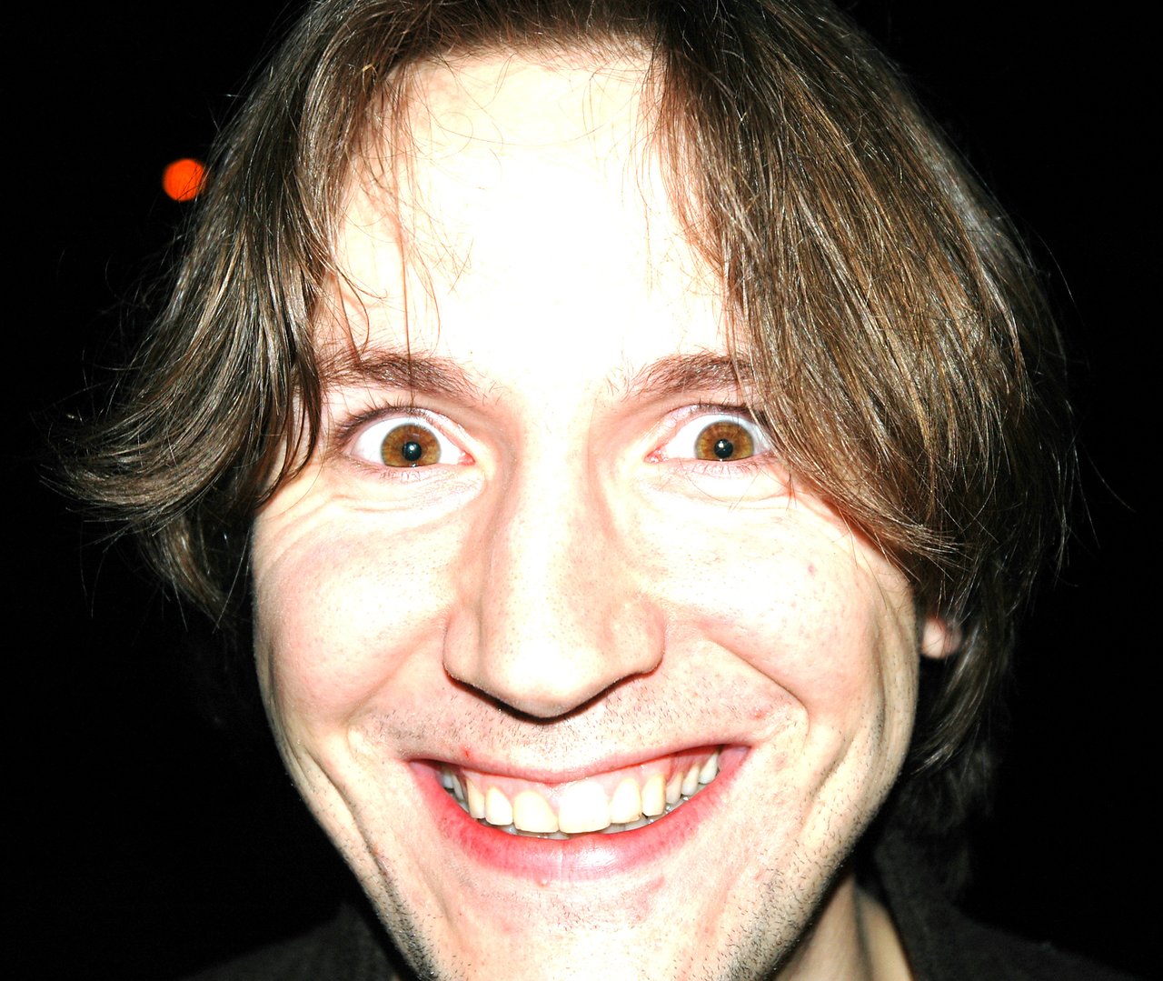 A person with light skin and medium-length hair smiles widely, looking directly at the camera with bright eyes.