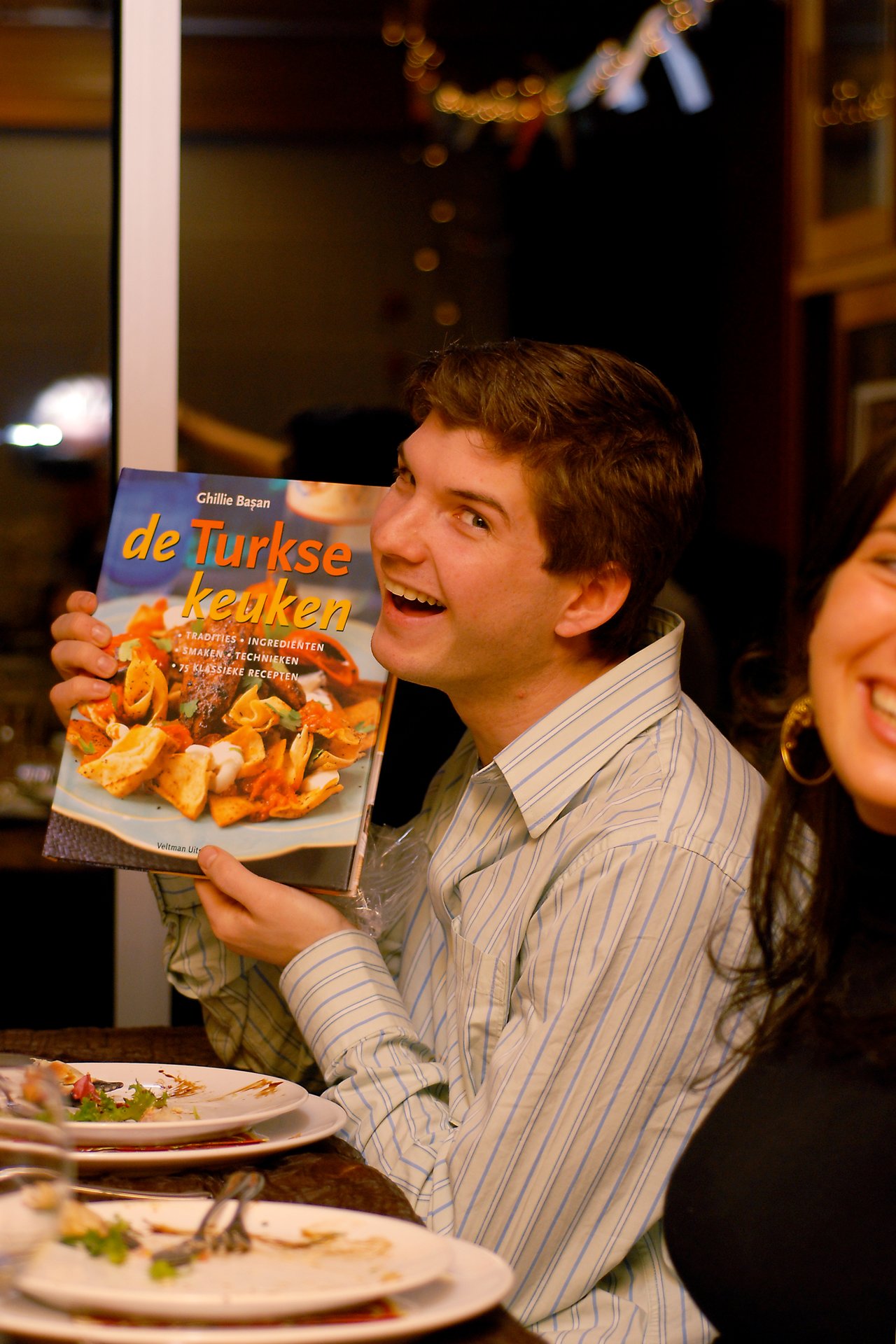 A smiling man holds up a Turkish cookbook at a dinner table, with a woman laughing beside him.