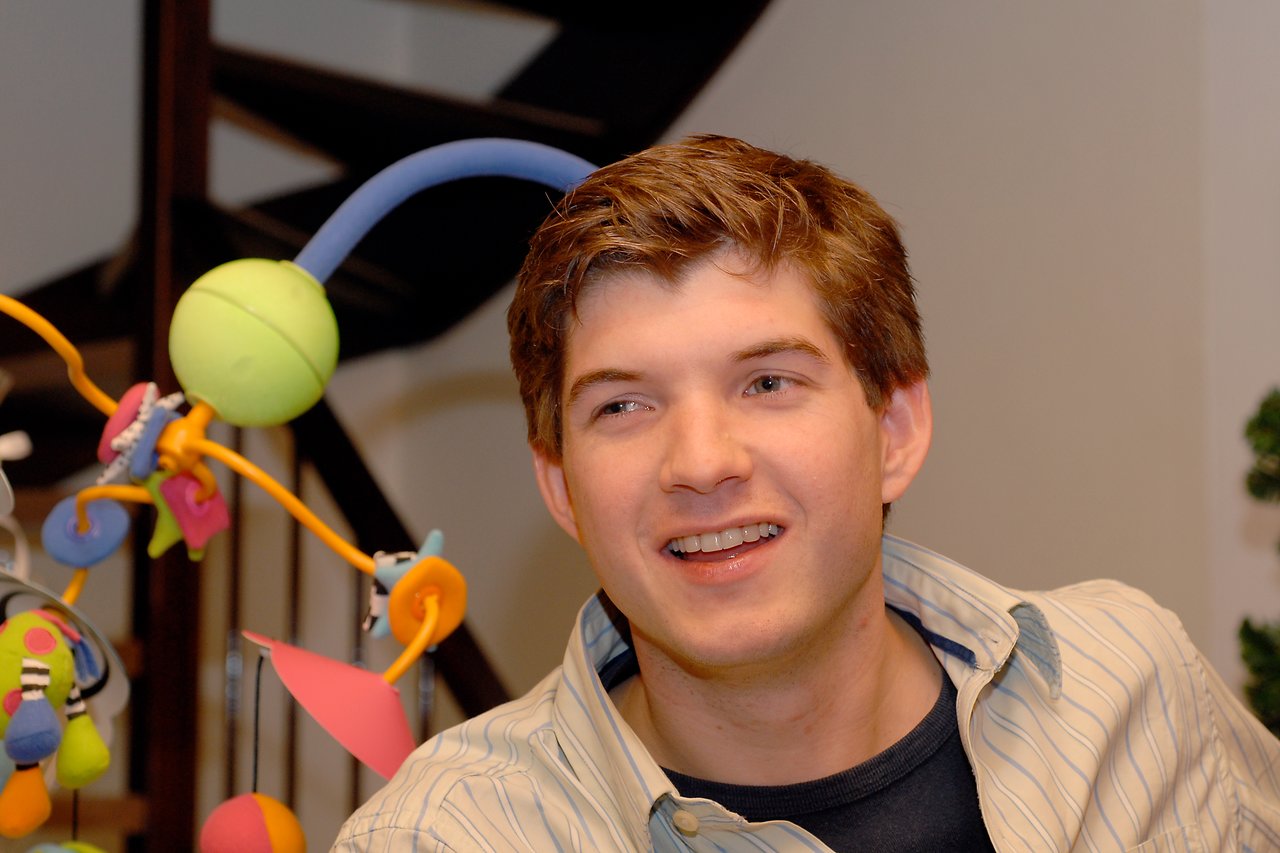A young man with short brown hair smiles while looking off to the side, sitting near a colorful toy structure.