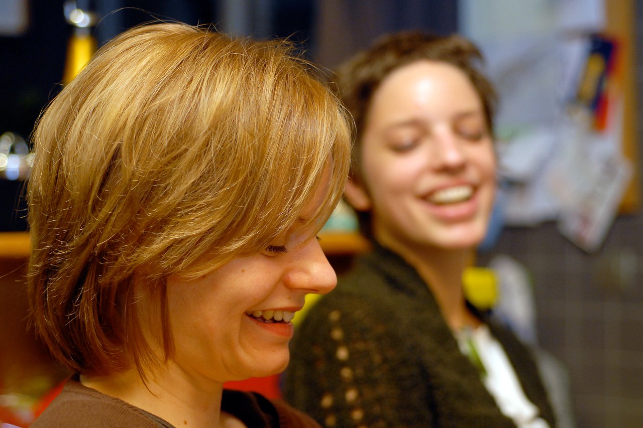 Two women laughing together, one in focus smiling while the other in the background is slightly blurred.