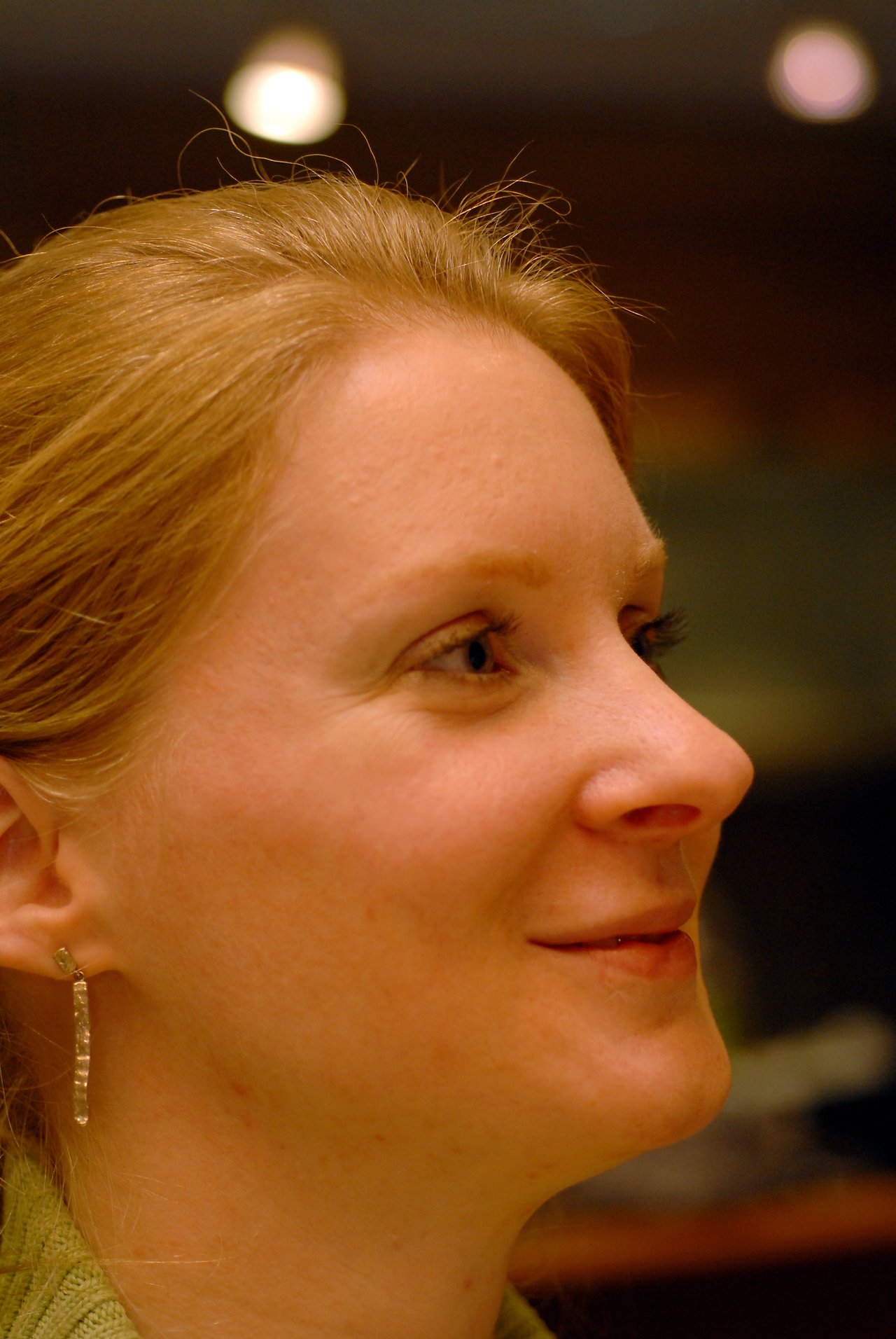 A woman with light skin and blonde hair smiles slightly while looking to the side in a warmly lit setting.