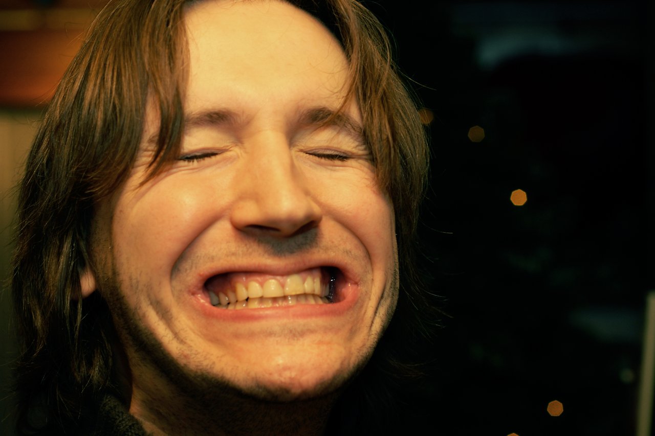 A person with long hair smiles widely with eyes closed, showing teeth in a playful expression.