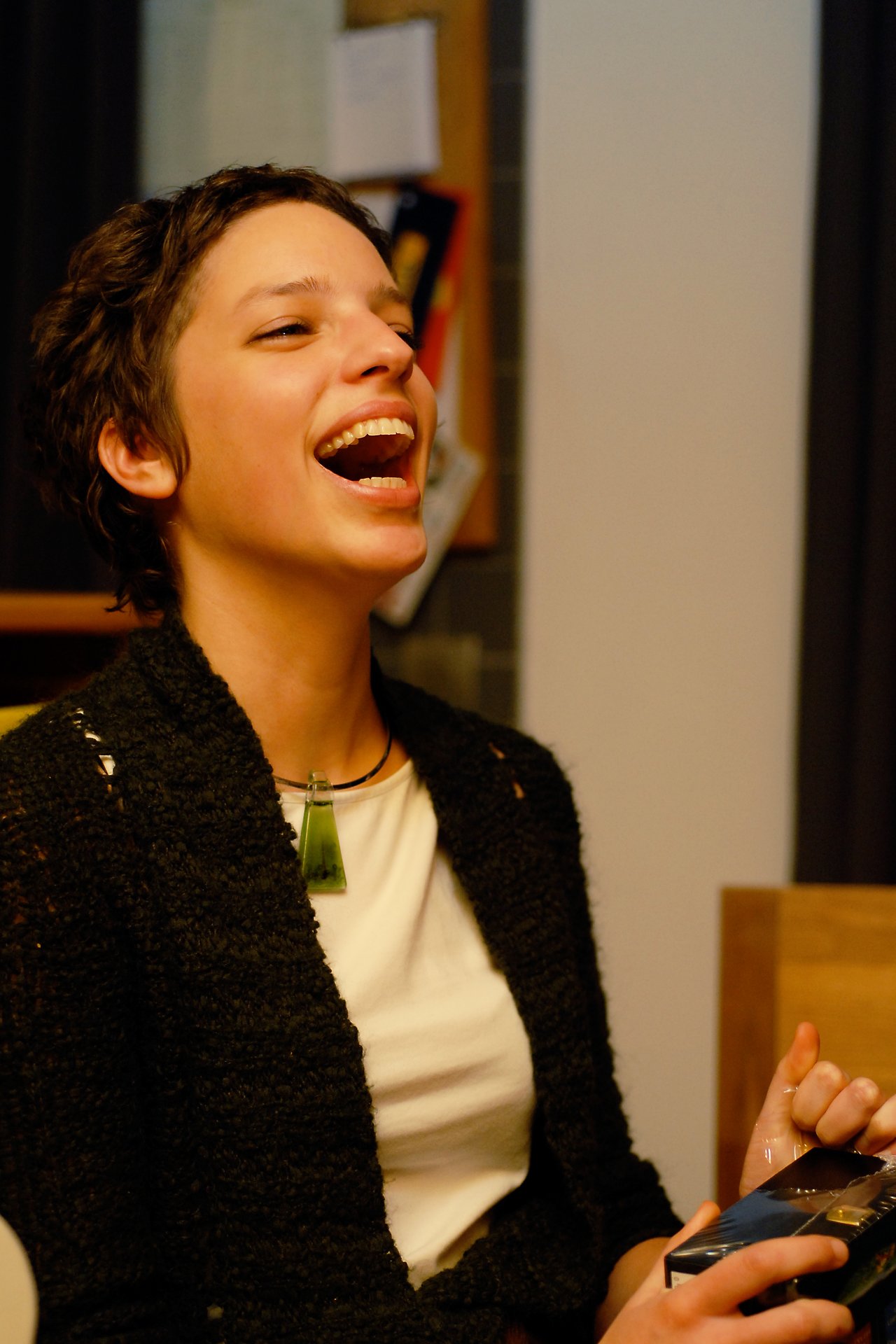 A person laughing while holding a small box, wearing a black sweater and a green pendant necklace.