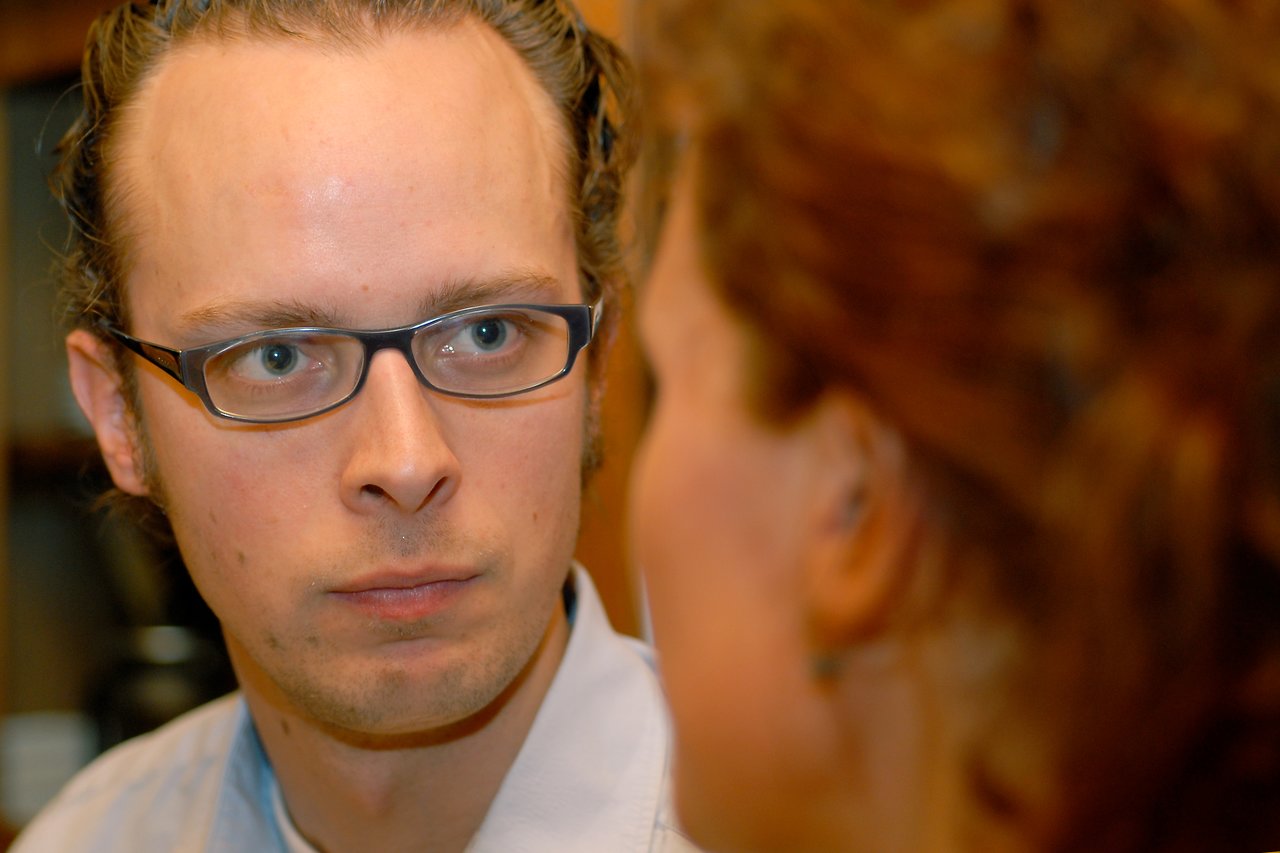 A man wearing glasses looks intently at a woman in the foreground during a conversation.
