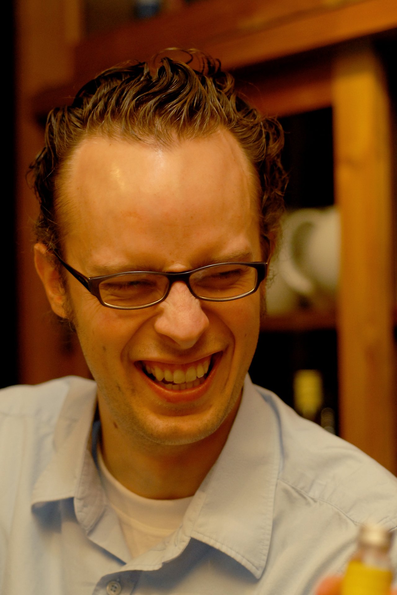 A man wearing glasses and a light blue shirt laughs with his eyes closed in a warmly lit room.
