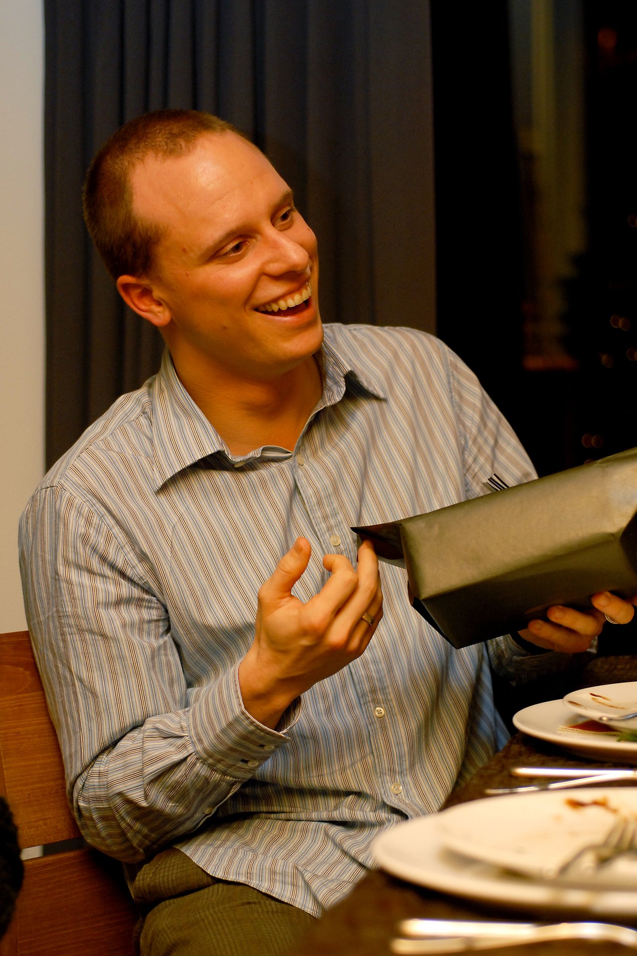 A smiling man in a striped shirt holds a wrapped gift while sitting at a dinner table.