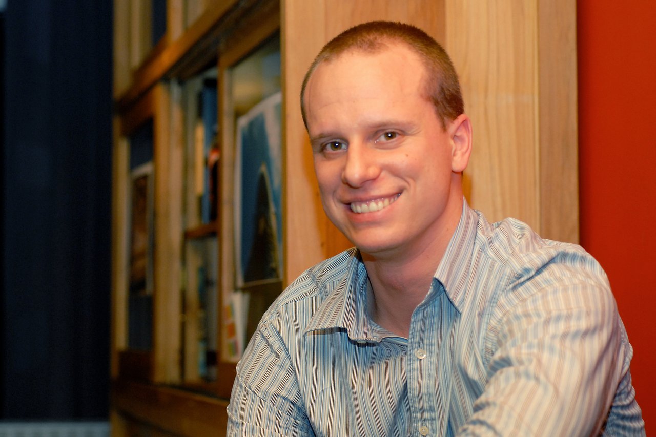 A man in a striped shirt smiles at the camera while sitting indoors near a wooden wall with framed pictures.