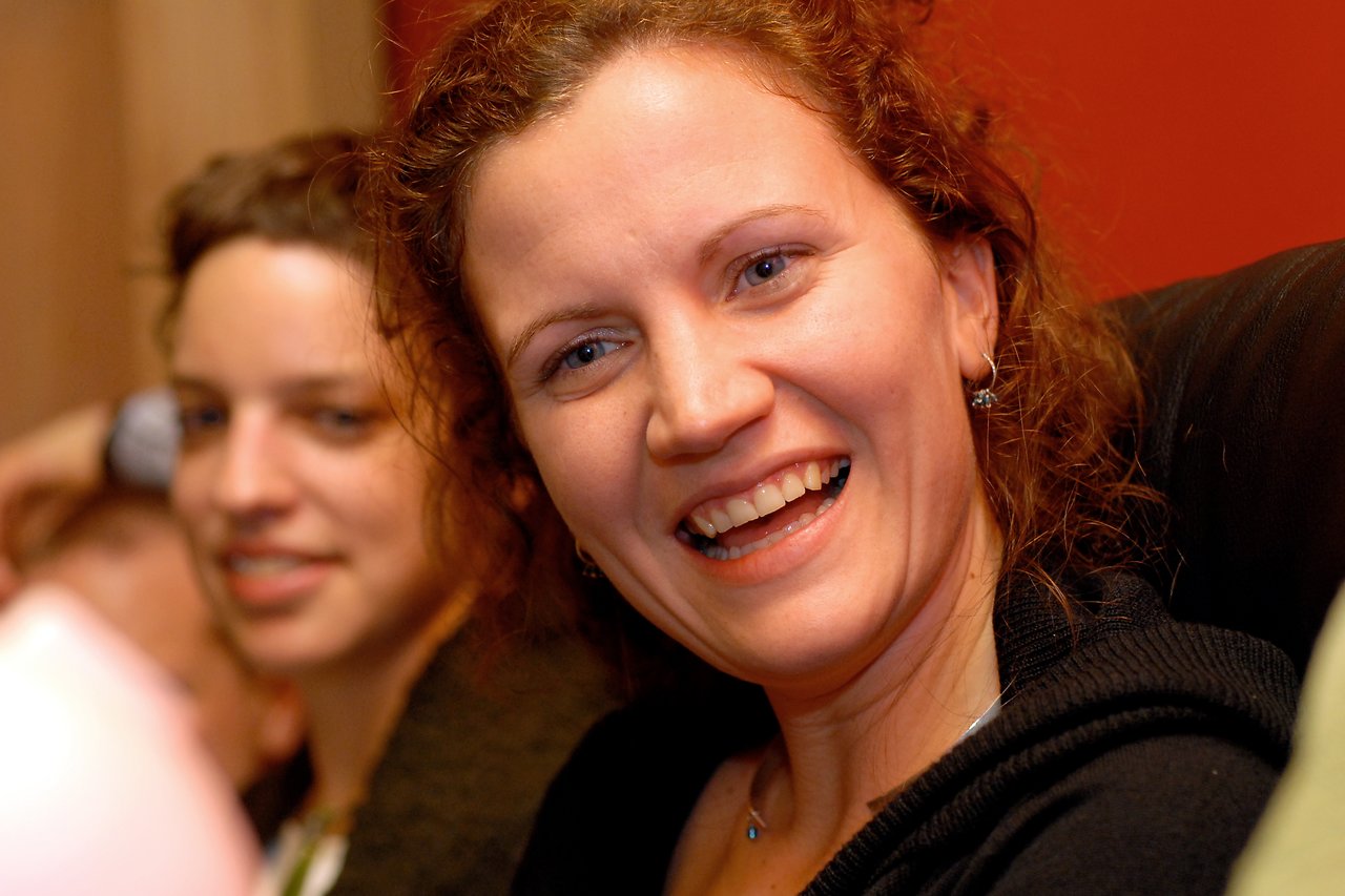 A woman with curly hair smiles brightly while sitting in a social setting, with another person slightly blurred behind her.