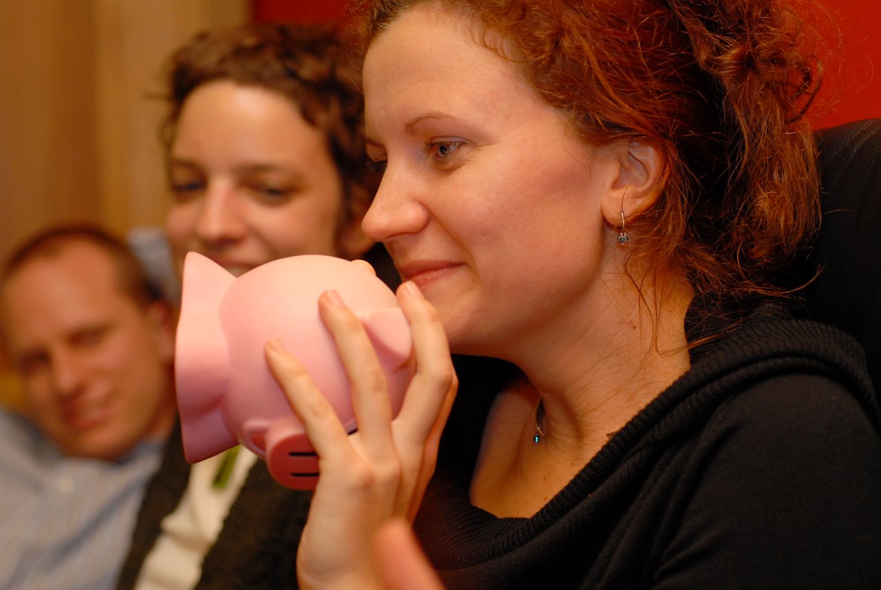 A woman holds a pink piggy bank close to her face while smiling, with two people watching in the background.