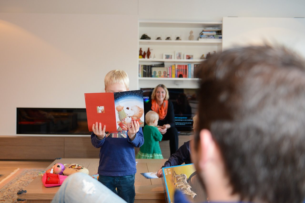 A child in a blue shirt holds up a holiday card, while others sit and interact in a living room.