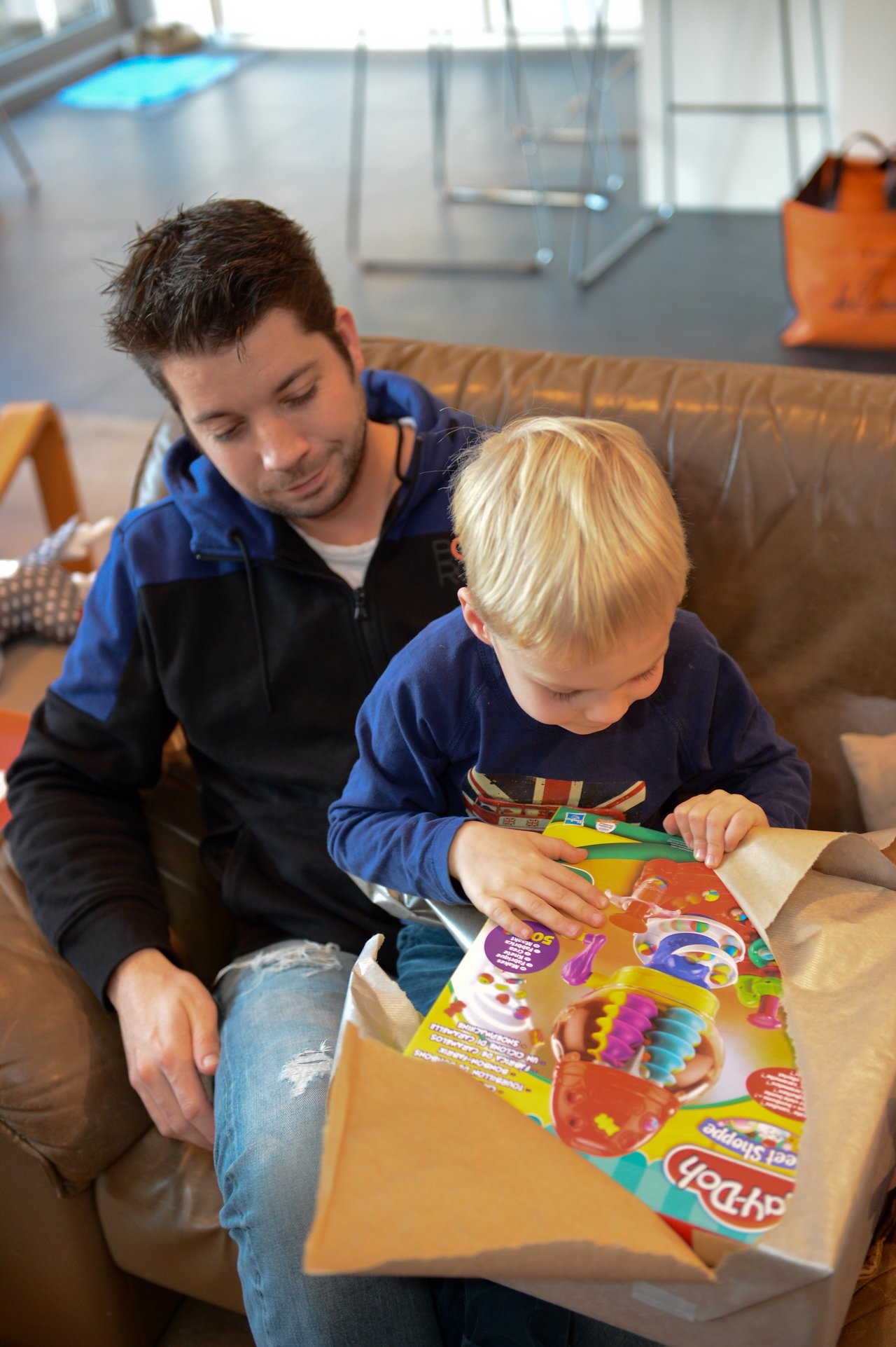 A child sits on an adult's lap, unwrapping a Play-Doh toy while looking at it with interest.