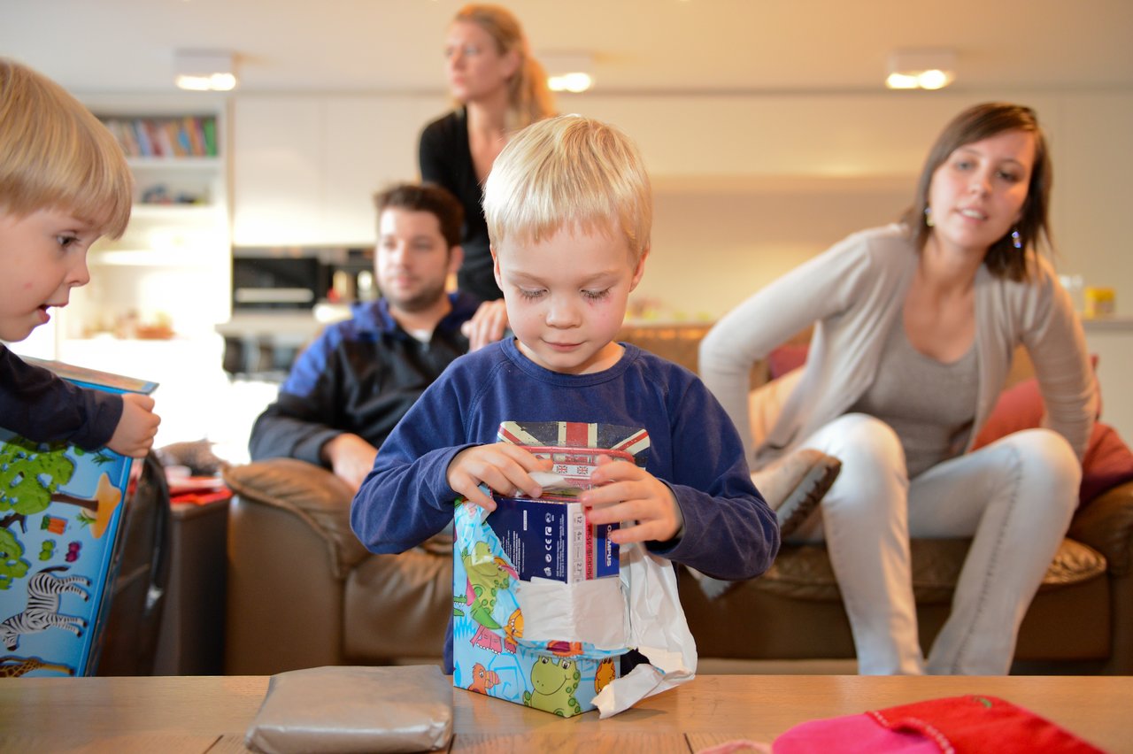 A young child opens a gift while another child watches, with adults sitting in the background.