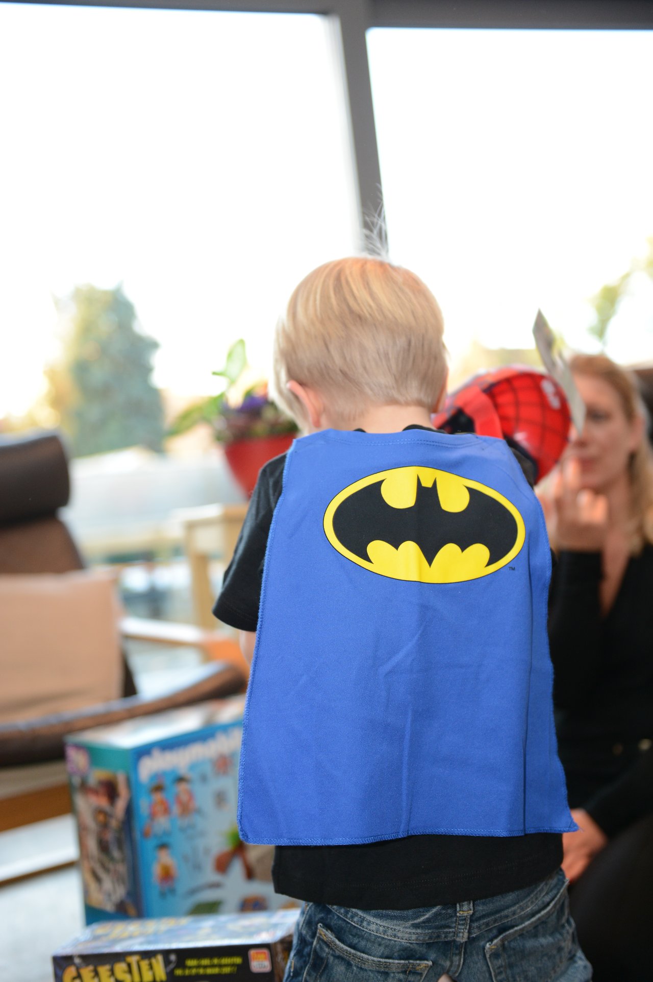 A child wearing a Batman cape stands near toys and decorations at a party.