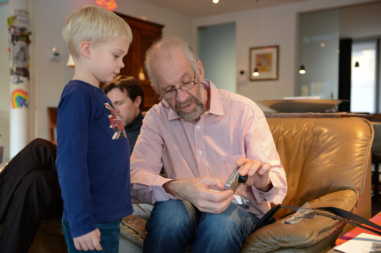 An older man sits on a couch, opening a package while a young boy watches closely.