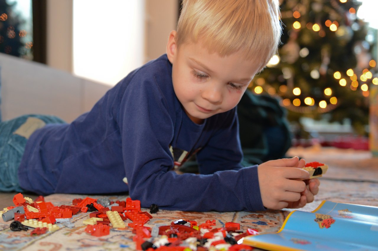 A young child in a blue shirt builds with LEGO bricks while lying on the floor near a Christmas tree.