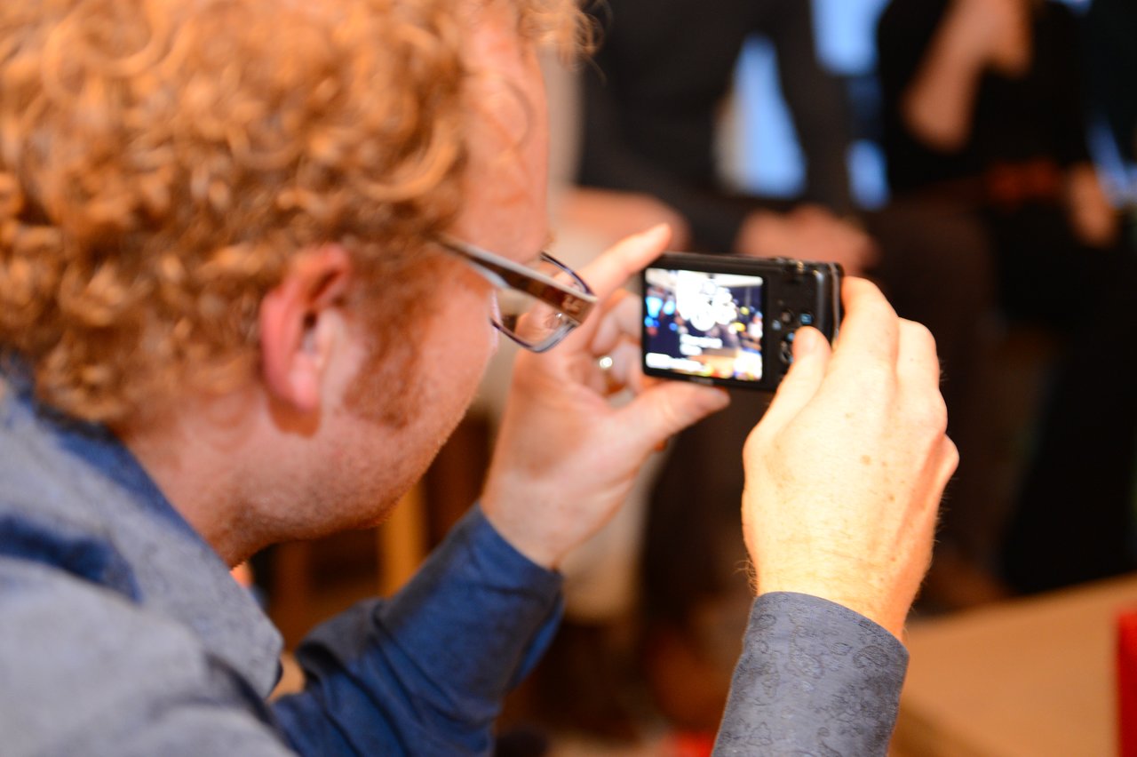 A person with curly hair and glasses takes a photo with a small camera at a party.