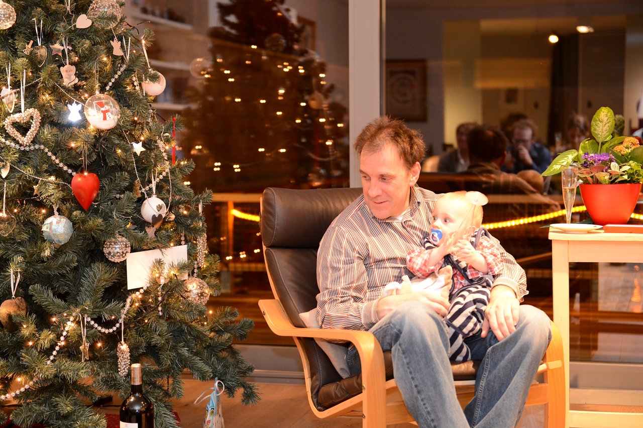 A man sits in a chair holding a baby, next to a decorated Christmas tree at a festive gathering.