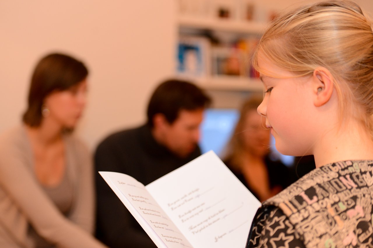 A young girl reads from a card while a group of adults sits in the background.
