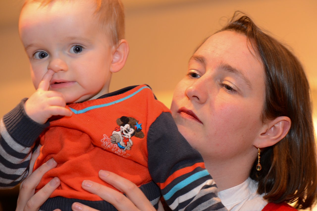 A woman holds a baby wearing an orange sweater, who is touching their nose with a finger.