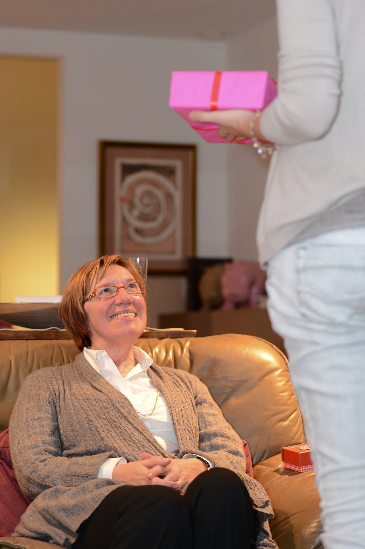 A seated woman smiles as she looks up at someone handing her a pink gift box.