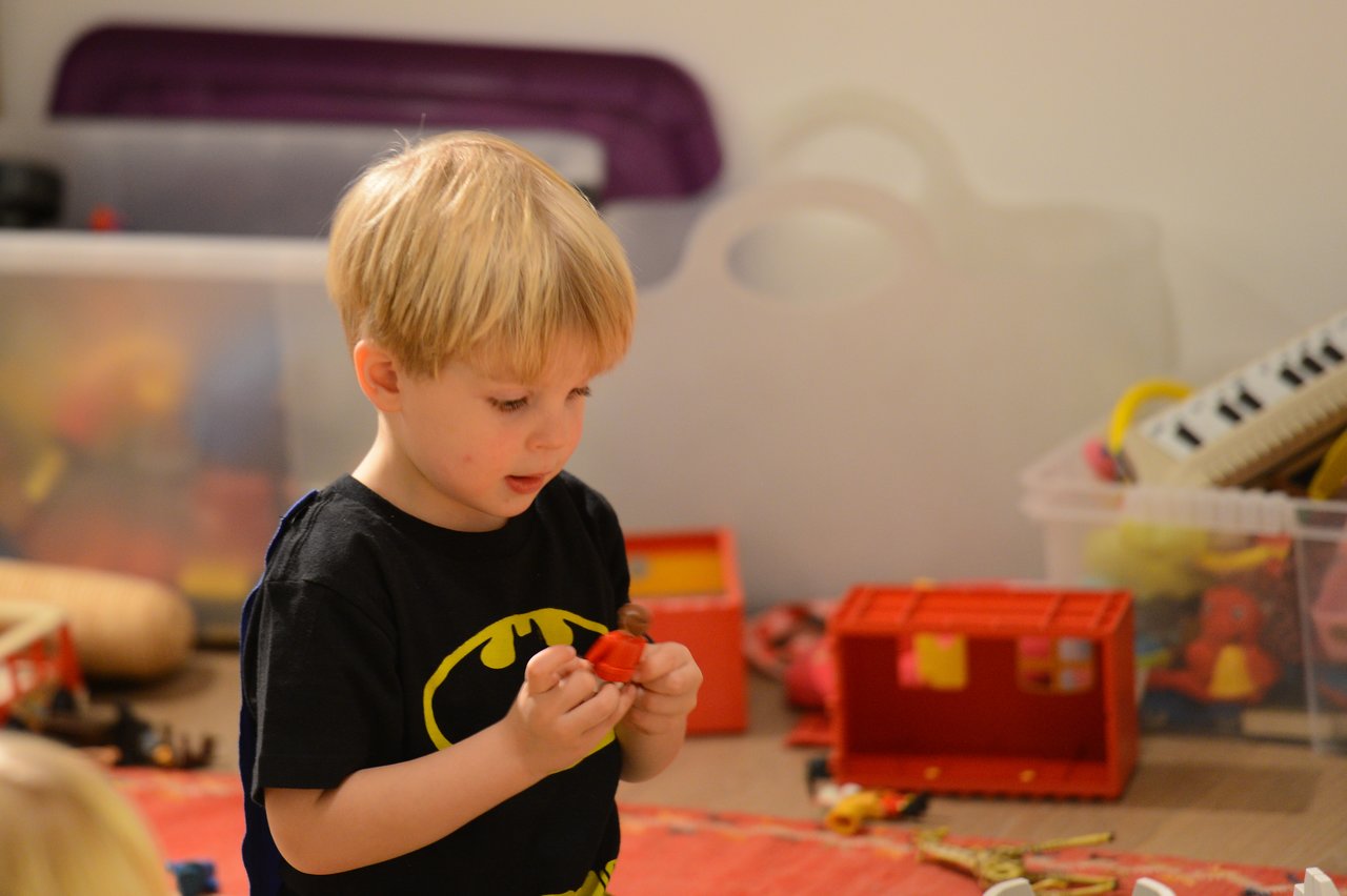 A young child in a Batman shirt holds a small toy while playing in a room with scattered toys.