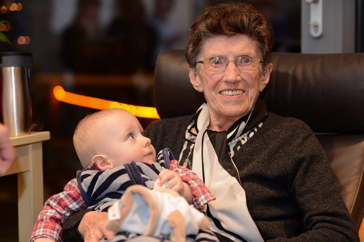 An elderly woman smiles while holding a baby who looks up at her during a gathering.