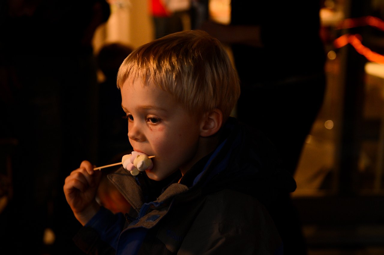 A young child eats marshmallows on a stick at a party, wearing a jacket in a dimly lit setting.