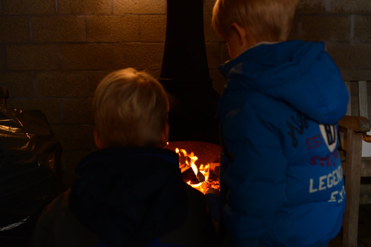 Two children in blue jackets stand near a fireplace, watching the flames.