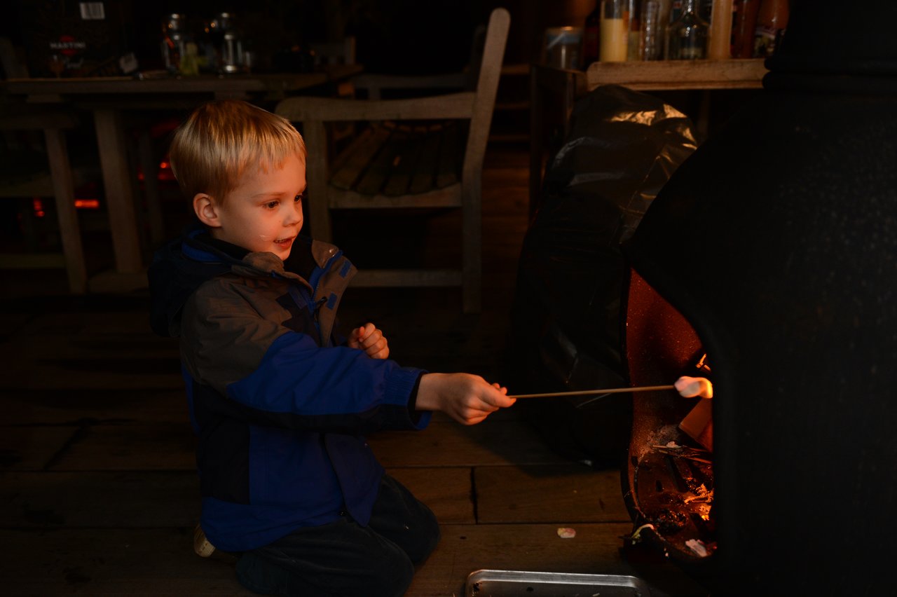 A young child in a blue jacket roasts marshmallows over a fire at an indoor gathering.