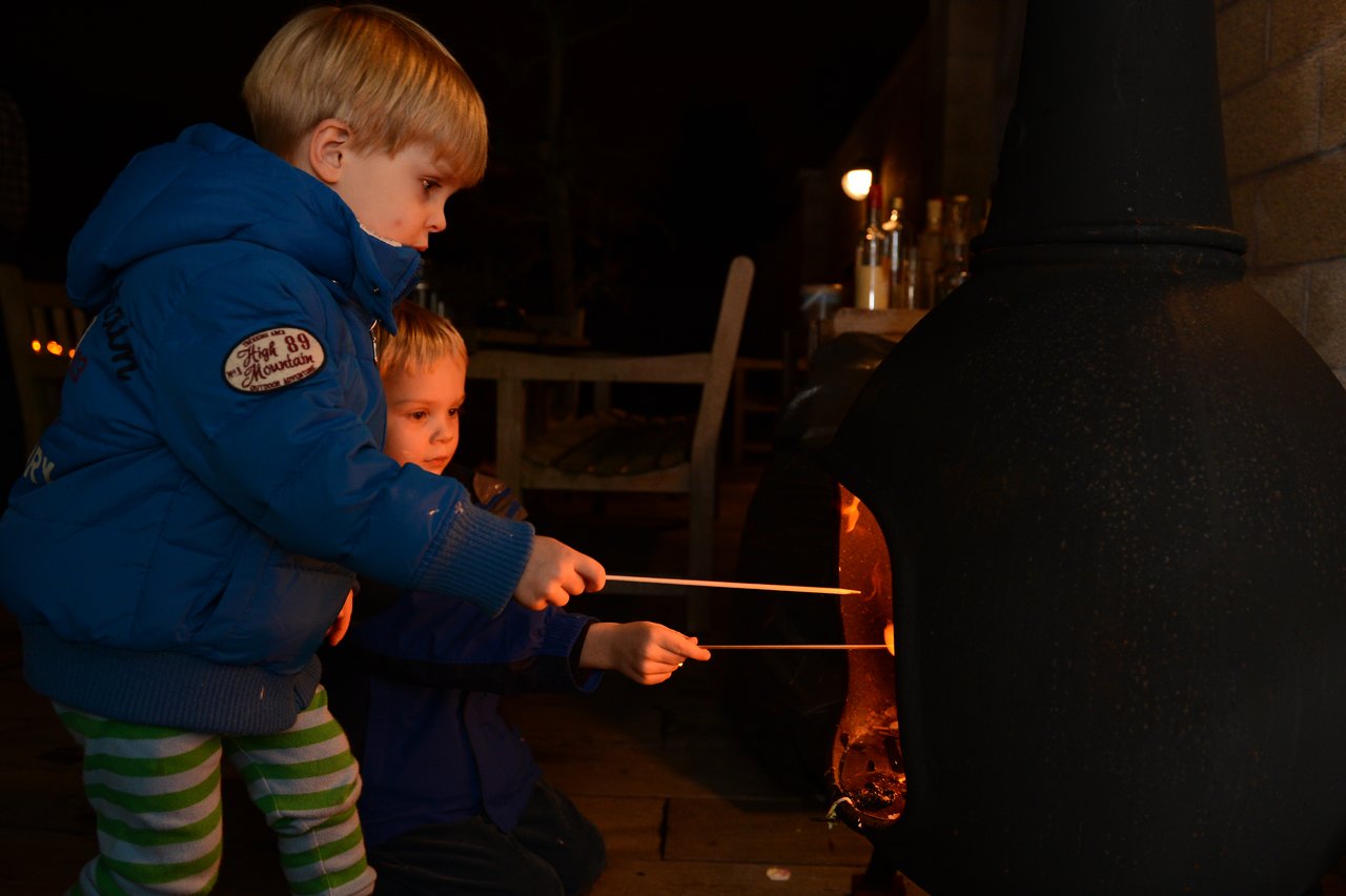 Two children in warm jackets roast marshmallows over an outdoor fire in a chiminea at night.